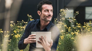 Person sitzt im Freien inmitten gelber Blüten und hält ein Tablet und einen Becher, während im Hintergrund dunkle Gebäudeteile sichtbar sind.