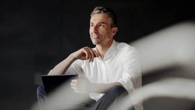 Person in hellem Hemd sitzt vor dunklem Hintergrund und hält ein Tablet in der Hand; die andere Hand ist nachdenklich am Kinn positioniert. Weiches Licht fällt seitlich ins Bild und erzeugt eine ruhige, konzentrierte Atmosphäre.