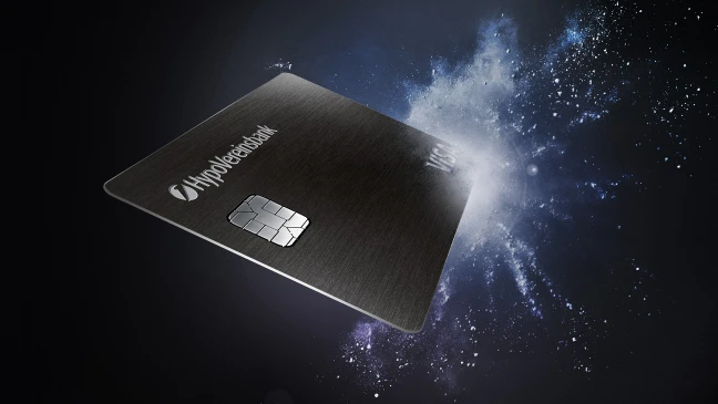 Schwarze HypoVereinsbank‑Kreditkarte mit silbernem Chip schwebt vor dunklem Hintergrund mit dynamischem Licht‑ und Partikeleffekt, der eine kraftvolle, moderne Anmutung erzeugt.