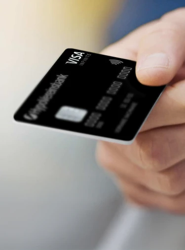 Hand hält eine schwarze Kreditkarte mit Logo HypoVereinsbank, Schriftzug VISA Infinite, Chip, Kontaktlos-Symbol und Zahlenreihe vor unscharfem Hintergrund.