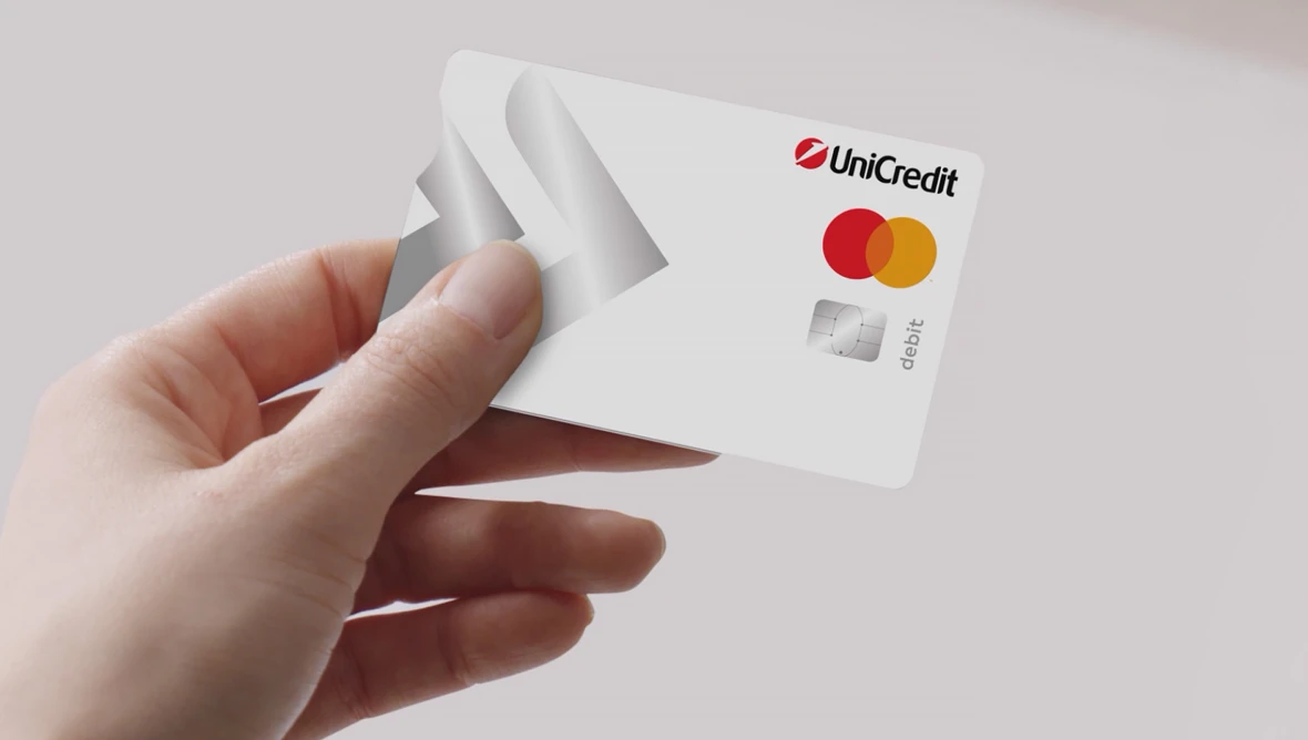 Hand hält weiße Debitkarte mit UniCredit Logo sowie Mastercard Zeichen und Chip vor neutralem Hintergrund mit grauem grafischem Element.