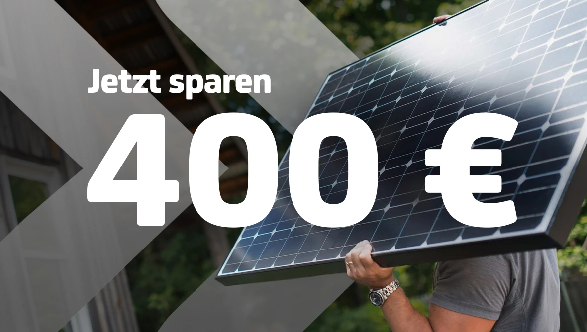 Person trägt Solarpanel, Text „Jetzt sparen 400 €“ im Vordergrund, unscharfer Hintergrund mit Gebäude und Bäumen.