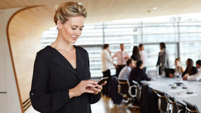 Blonde Frau mit lächelndem Gesichtsausdruck steht in einem Meetingraum und blickt auf ein Handy in ihrer Hand. 