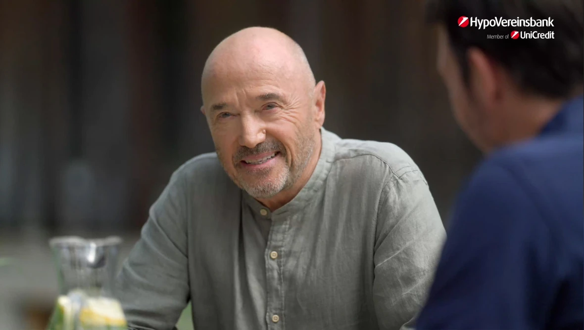 Christian und Felix Neureuther sitzen an einem Holztisch im Freien mit Wasserkrug und Gläsern, im Hintergrund dunkle Holzfläche, Logo oben rechts.