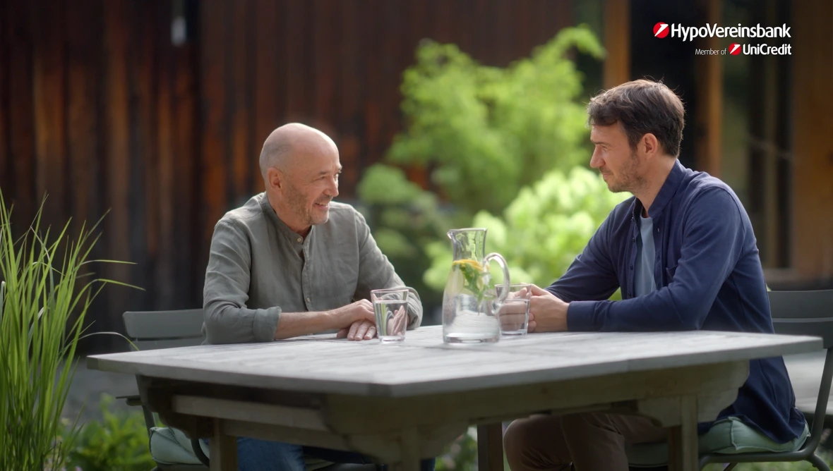 Christian und Felix Neureuther sitzen an einem Holztisch im Freien mit Wasserkrug und Gläsern, im Hintergrund Pflanzen, Logo oben rechts.
