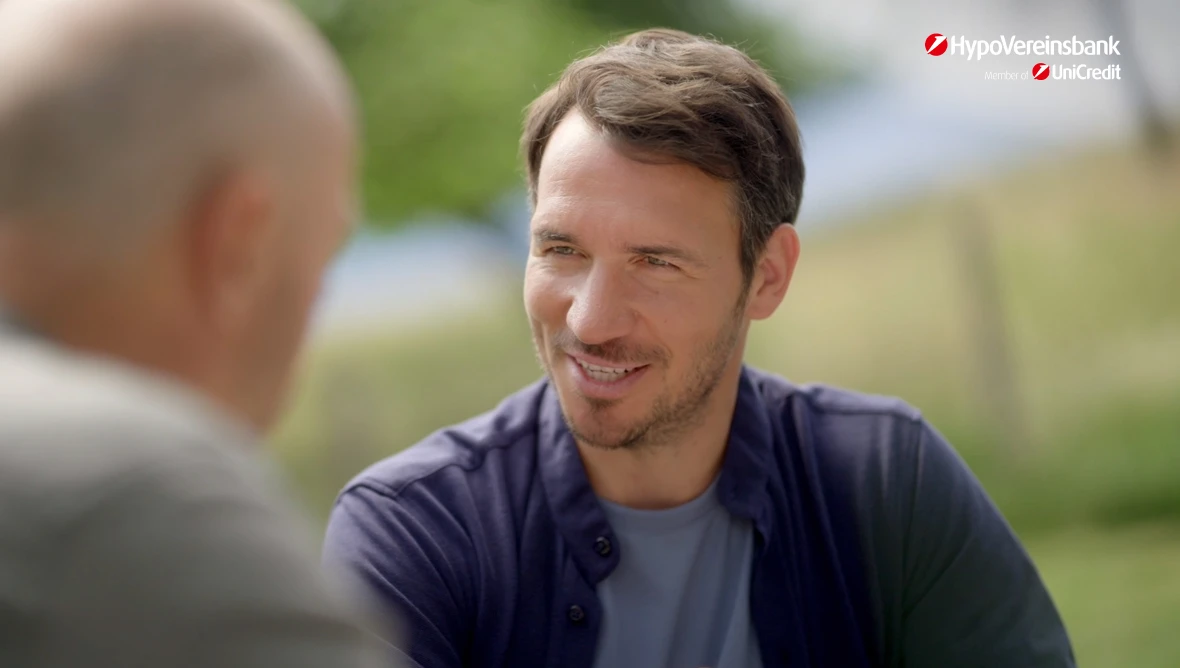 Felix und Christian Neureuther sitzen draußen an einem Tisch, im Hintergrund grüne Bäume und ein Zaun, oben rechts das HypoVereinsbank Logo.