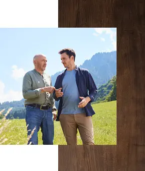 Christian und Felix Neureuther stehen auf einer grünen Wiese vor bewaldeten Hügeln und Bergen unter blauem Himmel, beide in Freizeitkleidung.