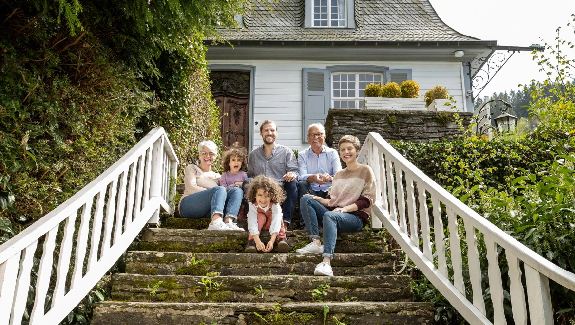 Mehrere Personen sitzen gemeinsam auf einer steinernen Außentreppe vor einem hellen Haus mit Fensterläden und Gartenpflanzen. Im Vordergrund sitzen zwei Kinder, dahinter weitere Personen. Weiße Geländer, begrünte Umgebung und Tageslicht prägen die Szene.