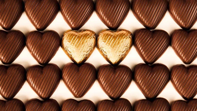 Reihe herzförmiger Pralinen, darunter zwei goldene Herzen als Blickfang zwischen dunkler Schokolade.