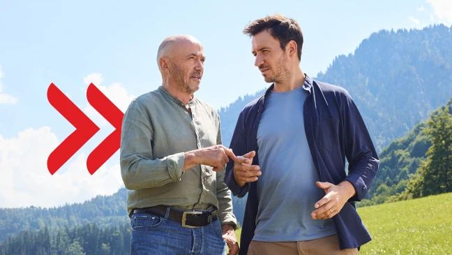 Zwei Personen (Christian und Felix Neureuther) stehen im Freien auf einer grünen Wiese vor einer Berglandschaft und unterhalten sich miteinander. Beide tragen legere Kleidung; links im Bild ist ein rotes, stilisiertes Pfeil Symbol zu sehen.