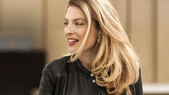 Person mit langen blonden Haaren und Lederjacke blickt lächelnd zur Seite, roter Lippenstift, unscharfer beiger Hintergrund.