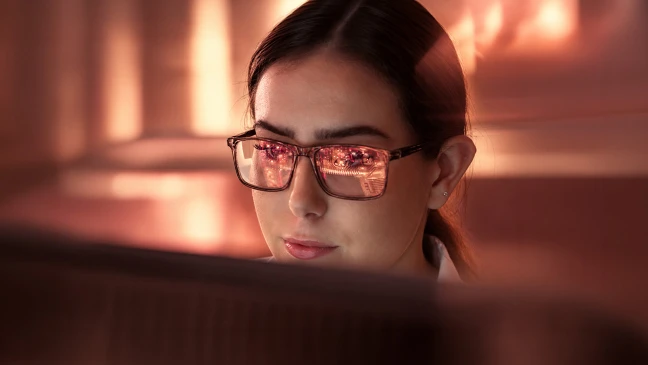 Person mit Brille blickt auf einen Bildschirm; Spiegelungen im Brillenglas unterstreichen digitales Arbeiten und Analyse.