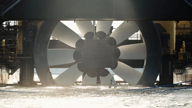 Große Turbine mit weißen Rotorblättern in technischer Anlage über Wasser, Person in Schutzkleidung steht davor im Wasser.