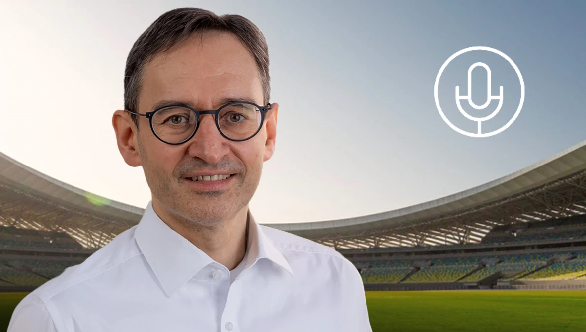 Person mit Brille und weißem Hemd vor Fußballstadion, rechts ein Mikrofon‑Symbol, Spielfeld und Tribünen im Hintergrund.