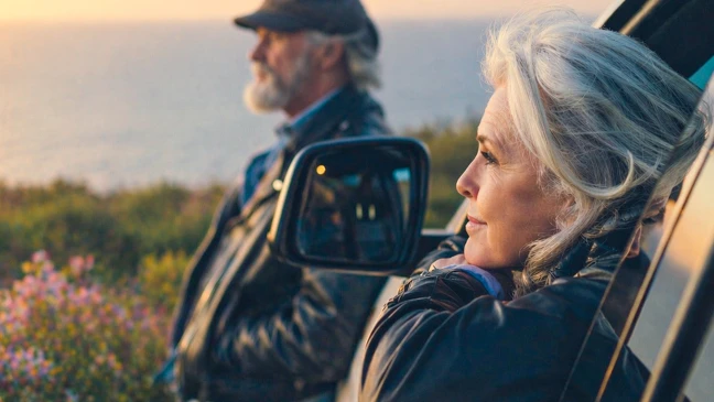 Zwei ältere Personen und ein geparkten Auto an der Küste. Eine Person lehnt sich aus dem offenen Autofenster, die andere steht daneben und blickt in die Ferne. Im Hintergrund sind Meer, Vegetation und warmes Abendlicht zu sehen.