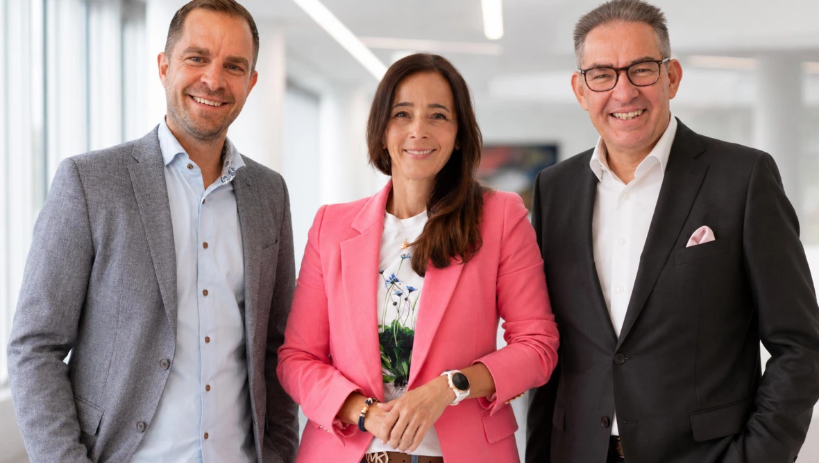 Drei Personen in Business-Kleidung stehen in einem hellen Büro und blicken in die Kamera.