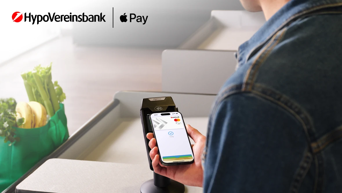 Person hält Smartphone an Kartenlesegerät neben Einkaufstasche, oben Logos von HVB und Apple Pay.