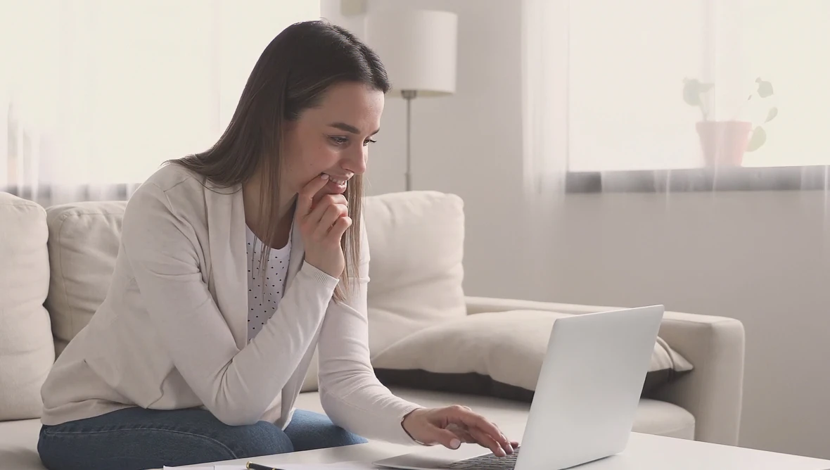 Person sitzt auf einem Sofa und arbeitet an einem aufgeklappten Laptop in einem hellen Raum mit Kissen und Pflanze im Hintergrund.