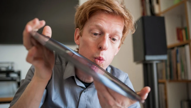 Eine Person hält ein aufgeklapptes Tablet oder Laptop in den Händen, im Innenraum mit Regalen und technischem Gerät im Hintergrund.