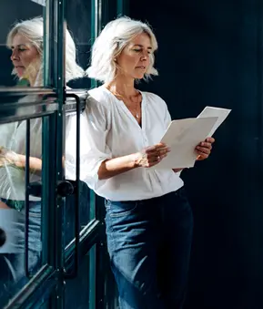 Eine Person lehnt an einer Glaswand und hält zwei Blätter Papier in den Händen, getragen werden eine helle Bluse und eine dunkle Hose.