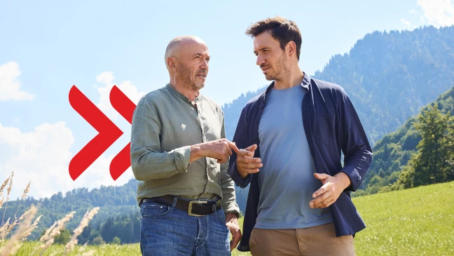 Christian und Felix Neureuther stehen auf einer Wiese vor einer Berglandschaft und unterhalten sich, während hinter ihnen ein großes rotes grafisches Symbol sichtbar ist.