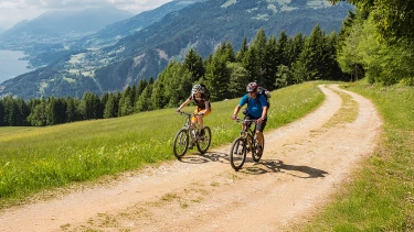 Zwei Personen fahren mit Fahrrädern auf einem kurvigen Schotterweg durch eine grüne Berglandschaft mit Wiesen und Nadelbäumen.