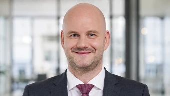 Portrait einer Person in dunklem Anzug und weißem Hemd mit roter Krawatte vor unscharfem Bürohintergrund.
