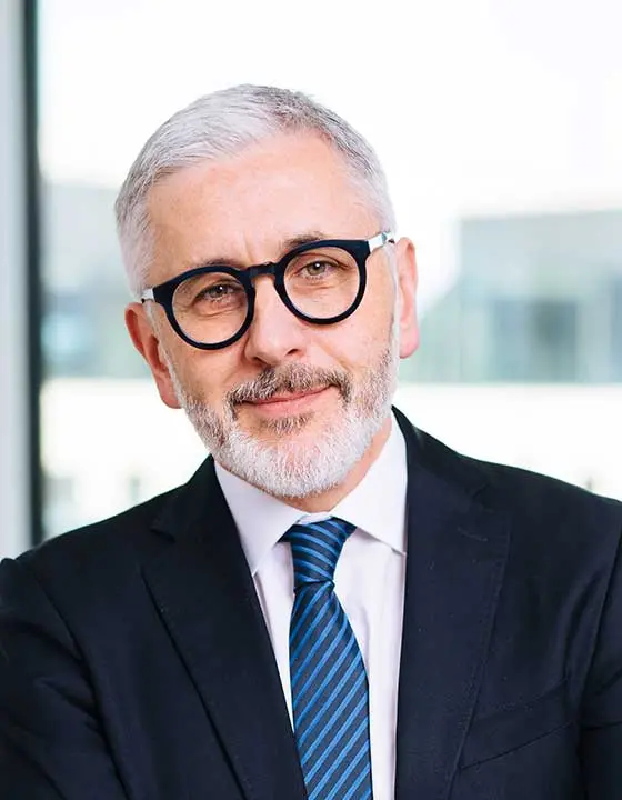 Portrait von Wolfgang Schilk mit grau melierten Haaren und einer Brille mit schwarzem Rahmen, in einem dunkelblauen Anzug mit blau gestreifter Krawatte und weißem Hemd, in einem modernen Bürogebäude.