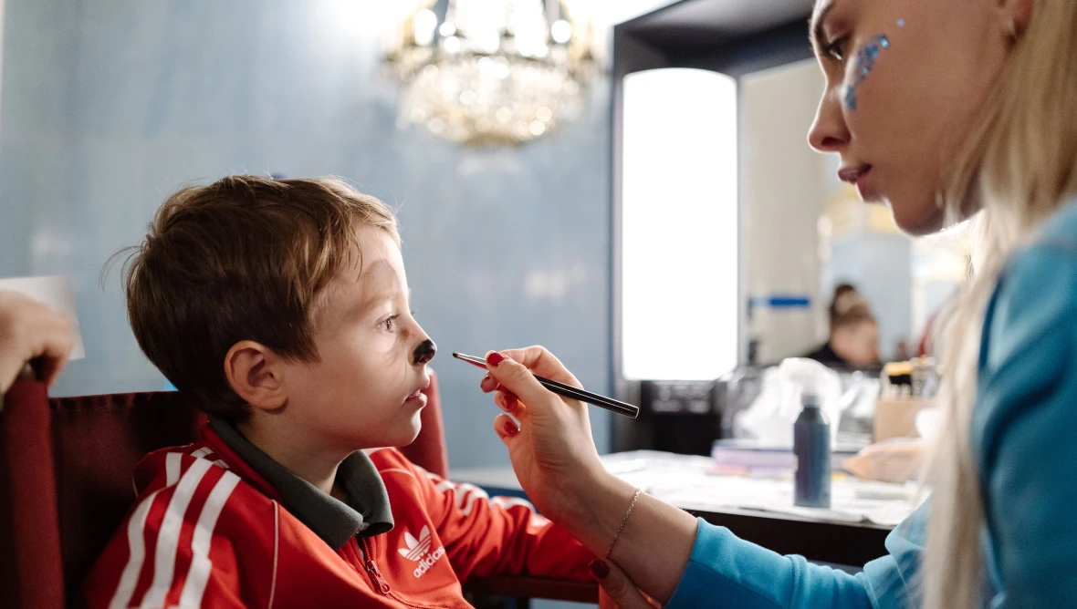 Ein Junge sitzt links auf einem Stuhl und wird gerade von einer Frau mit langen blonden Haaren, die rechts neben im sitzt, im Gesicht geschminkt
