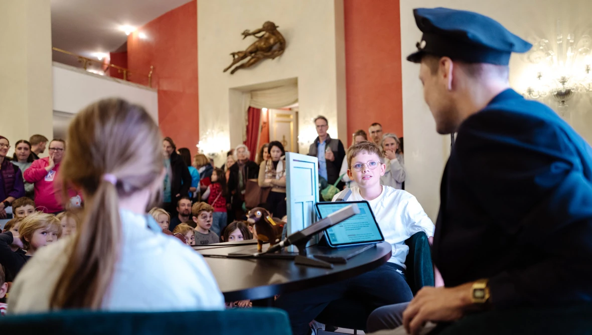 Man sieht ein Mädchen von hinten, rechts daneben sitzt ein Mann mit einer blauen Kappe, daneben ein Junge in einem weißen T-Shirt, vor ihnen steht ein Mikrophon und ein Tablet. Im Hintergrund sitzen und stehen Personen, die den dreien zusehen.
