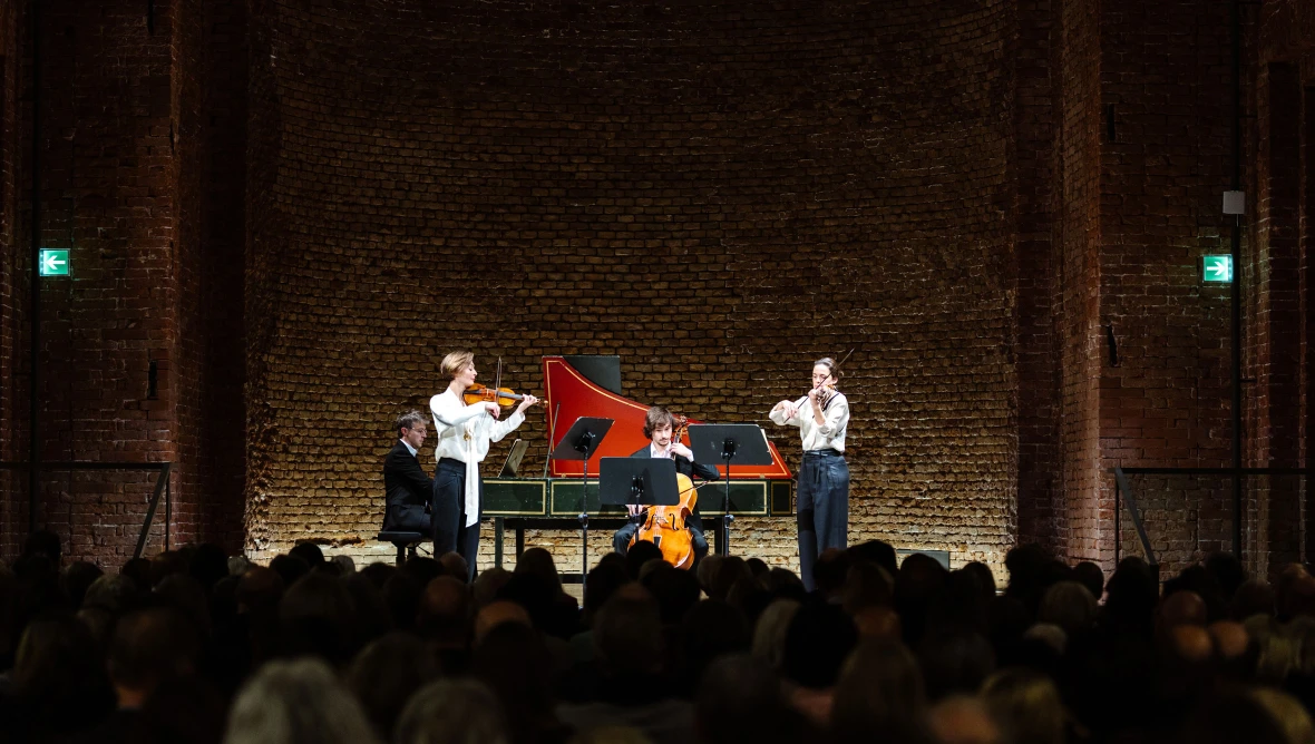 Eine Bühne auf der ein Pianist auf einem Flügel spielt, links davon spielt eine Frau ein Geige, in der Mitte spielt ein Mann Cello und rechts daneben spielt eine Frau Geige, davor ist im Dunklen das Publikum zu sehen, das vor der Bühne sitzt und zuhört.