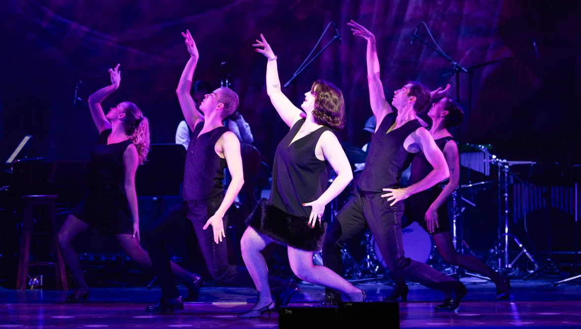 Auf der Bühne stehen sechs Tänzer:innen von blaumem Licht erstrahlt. Alle tragen schwarze Kleidung, haben die rechte Hand nach oben und haben das rechte Knie gebeugt.