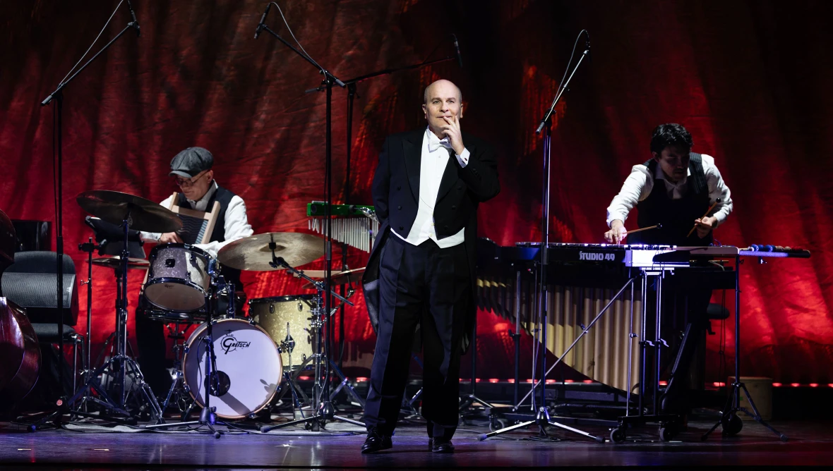 Auf der Bühne steht in der Mitte ein Sänger in einem schwarzen Frack, im Hintergrund spielen zwei Musiker am Schlagzeug und auf Schlaginstrumenten.