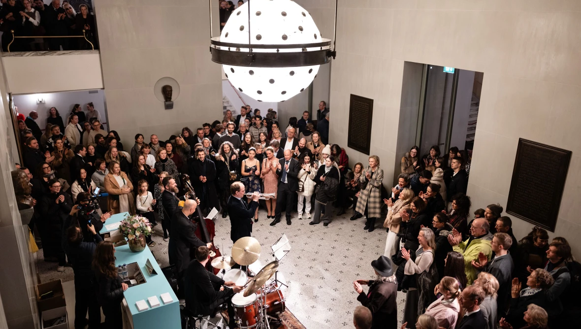 Man sieht von oben auf einen hellen Saal, es spielen vier Musiker Gitarre, Kontrabass, Schlagzeug und Trompete. Rundherum stehen Personen und klatschen.