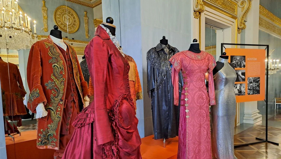 Fünf Kleiderbüsten die in einem barocken Saal mit Kronleuchter aufgstellt sind und unterschiedliche Kostüme (barocke Abendkleider, Pailettenkleider und Fracks) tragen.