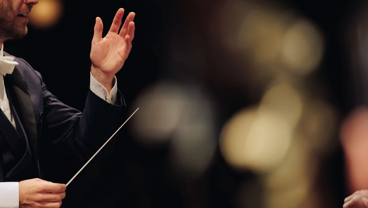 Nahaufnahme eines Dirigenten in einem schwarzen Frack und weißer Fliege am linken Bildrand, es ist ein Teil des Gesichtes zu sehen, der Mann streckt den linken Arm nach oben, in der rechten Hand hält er einen Taktstock. Der Hintergrund ist nur ganz unscharf zu sehen. 