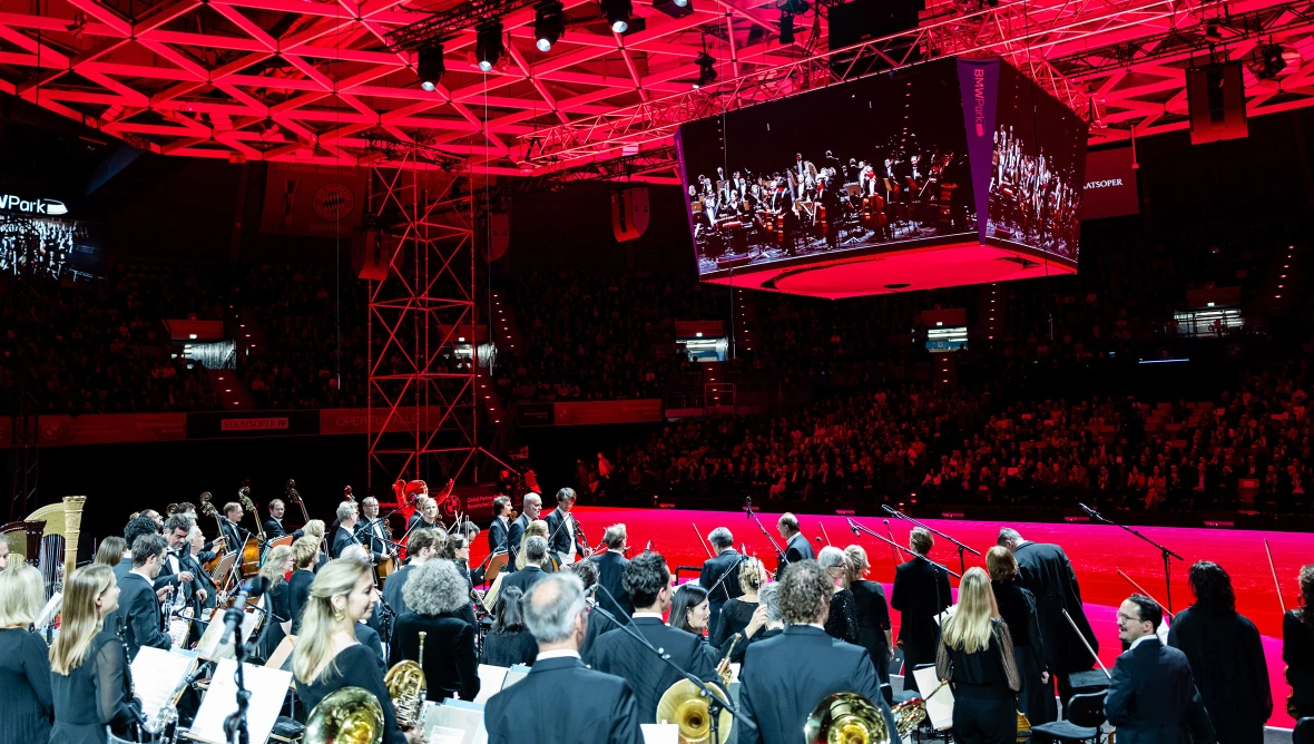 Mitglieder eines Orchesters stehen mit ihren Instrumenten in der Hand auf einer Bühne und blicken auf die Zuschauerränge, die Veranstaltungslocation ist in rotes Scheinwerferlicht getaucht