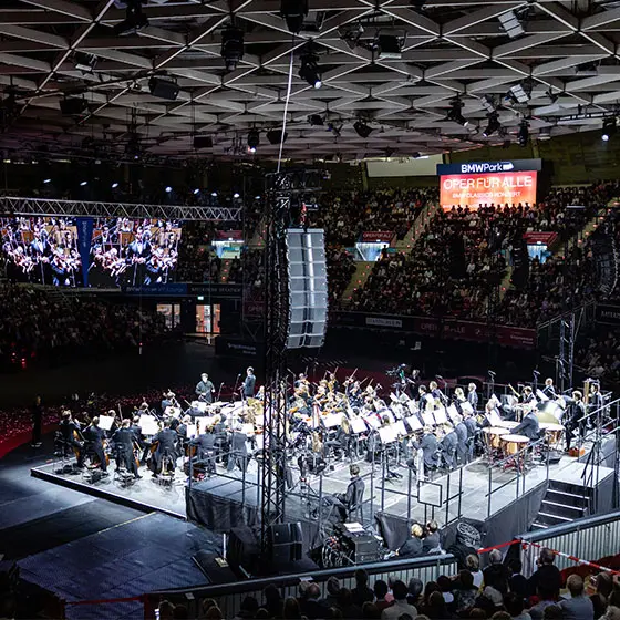 Blick von oben auf eine Konzert-Bühne mit Orchester, rundherum voll besetzte Besucherränge. 