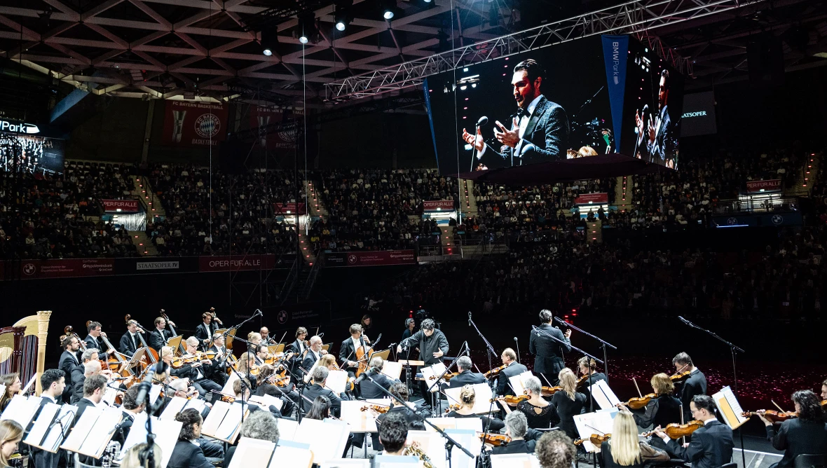 Blick von hinten auf die Bühne mit dem Orchester, rundherum die voll besetzten Besucherränge, oben sieht man einen Bildschirm, auf dem ein Opernsänger in Nahaufnahme zu sehen ist