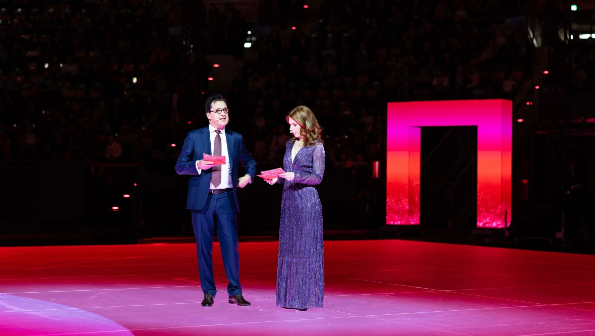 Ein Herr in einem dunkelblauen Anzug mit Krawatte und schwarzer Brille, rechts daneben steht eine Dame in einem dunkelblauem langem Abendkleid und blickt auf ihre Moderationskarten.