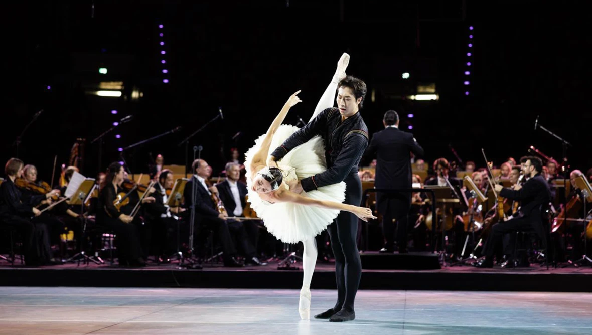 Eine Ballerina und ein Ballettänzer tanzen auf der Bühne, im Hintergrund ist ein Orchester zu sehen. 