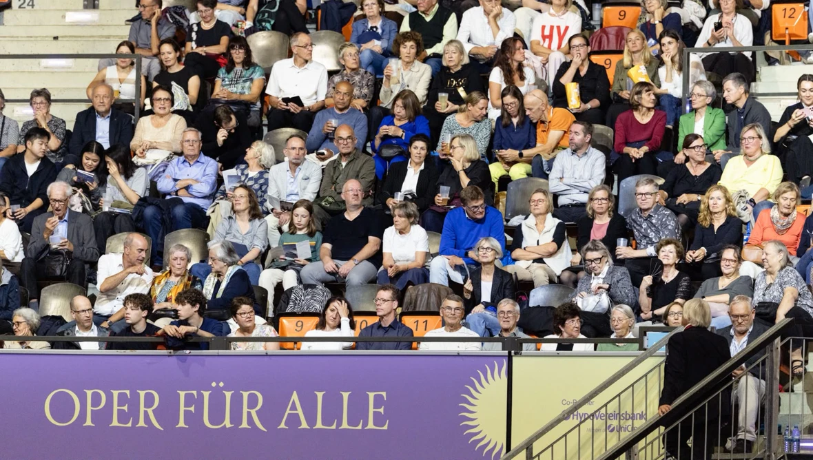 Zahlreiche Personen sitzen auf Zuschauerreihen über einem Banner mit der Aufschrift OPER FÜR ALLE.