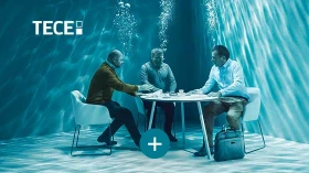 Drei Personen in formeller Kleidung sitzen an einem Tisch in einem mit Wasser gefüllten Raum, Blasen steigen auf.