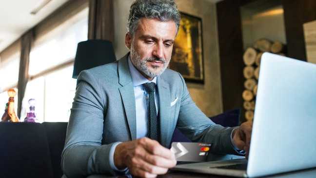 Person im Business‑Outfit arbeitet am Laptop und hält eine Kreditkarte in der Hand; modernes Wohnumfeld.