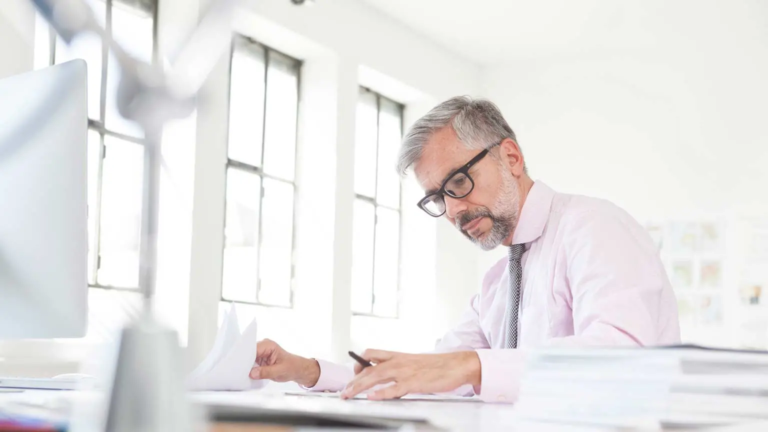 Eine Person mit Brille arbeitet in heller Umgebung an einem Schreibtisch mit Monitor und mehreren Papierstapeln..