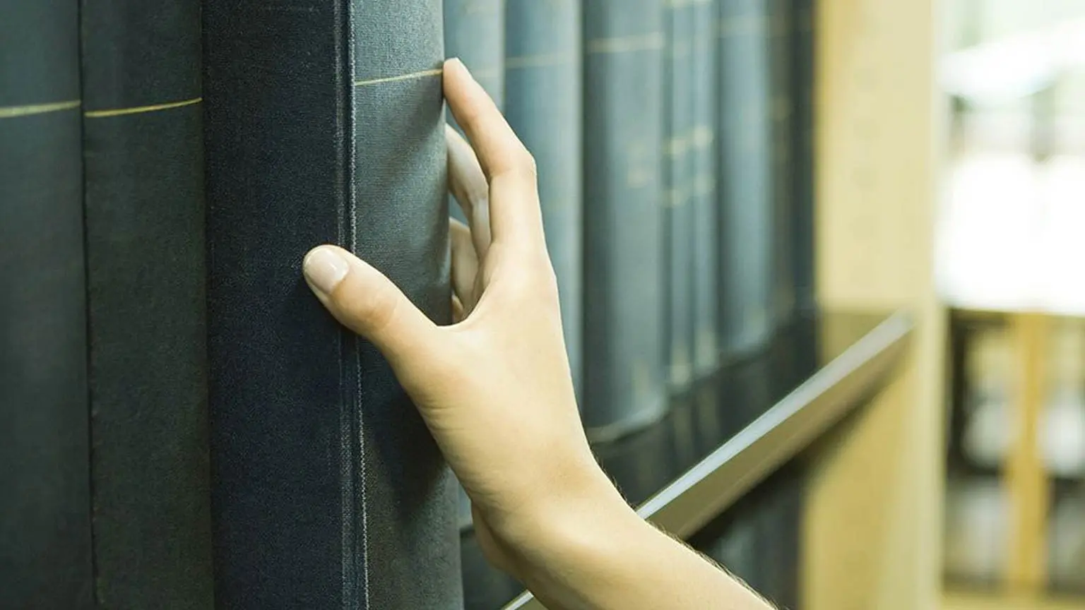 Eine Hand greift in einem Regal nach einem dicken, dunkel gebundenen Buch. Im Hintergrund ein großes Regal mit vielen Büchern.