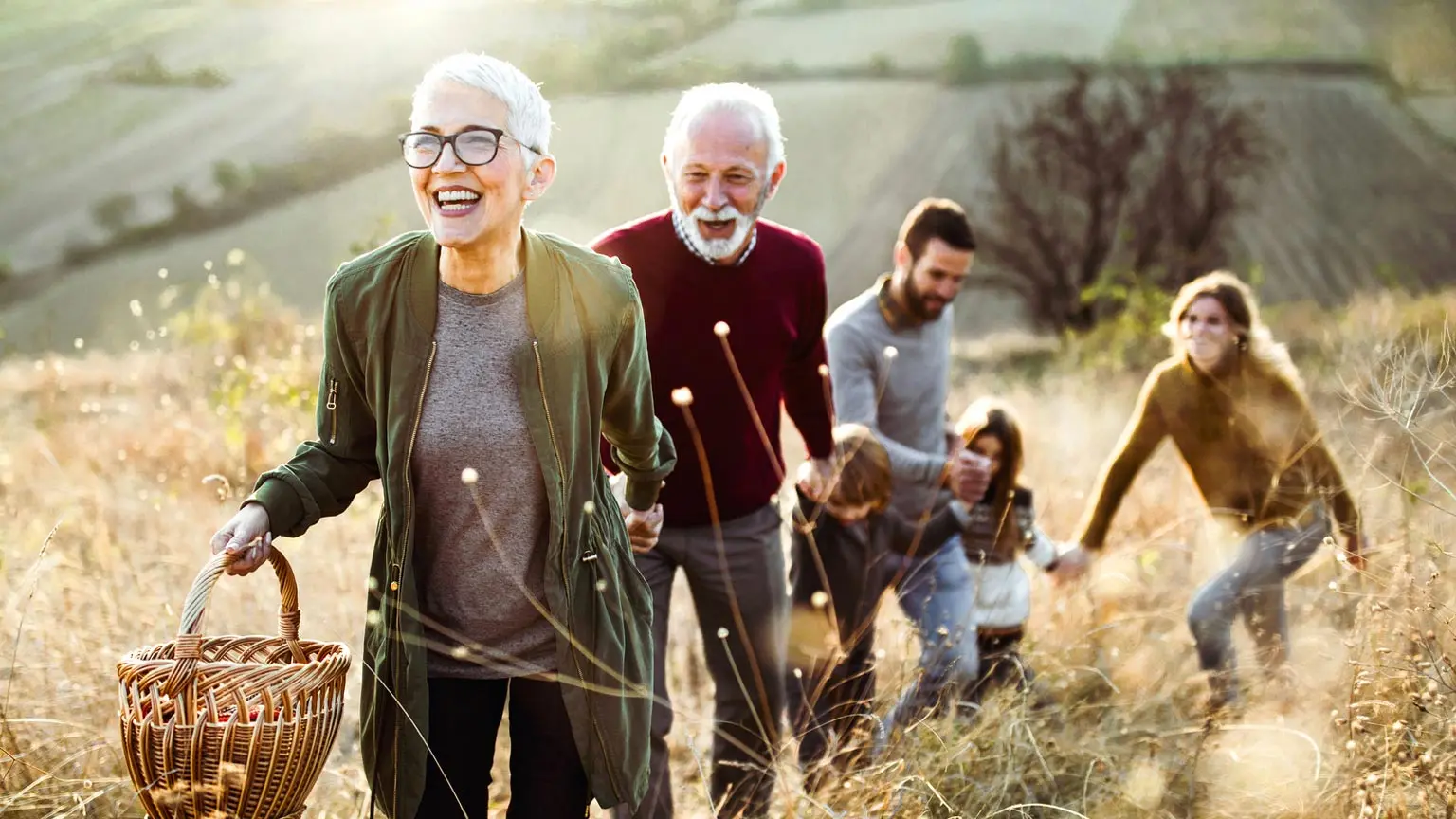 Gruppe von fünf Personen geht gemeinsam durch eine hügelige Landschaft mit trockenem Gras. Eine Person im Vordergrund trägt eine Brille und hält einen Korb, dahinter folgen weitere Personen, darunter auch zwei Kinder. Im Hintergrund sind Felder und sanfte Hügel zu sehen.