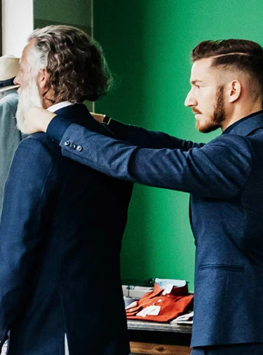 Zwei Personen in dunklen Anzügen, eine legt Hände auf die Schultern der anderen, im Raum mit grüner Wand, Tür, Kleiderständer und Tisch mit roten Stoffstücken.