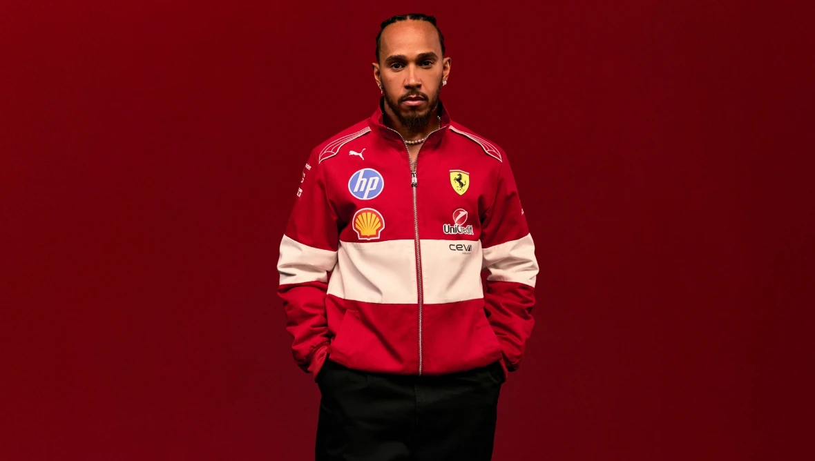Person trägt eine rote Ferrari-Teamjacke mit Sponsorenlogos wie Shell und HP, vor einem einfarbig roten Hintergrund.