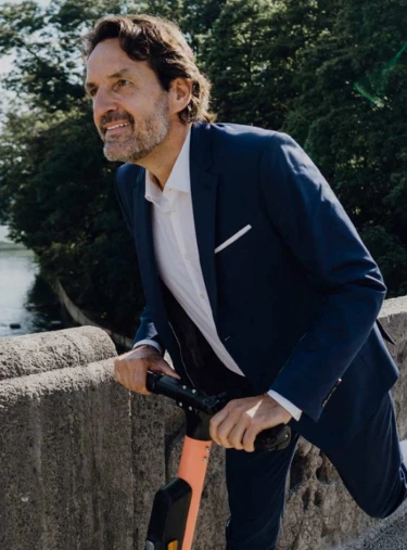 Person in dunkelblauem Anzug fährt auf einem Elektroroller über eine steinerne Brücke, im Hintergrund ein Fluss und grüne Bäume.